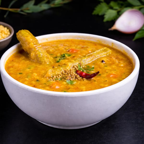 A white bowl filled with yellow dal, garnished with mustard seeds, red chili, and drumsticks.
